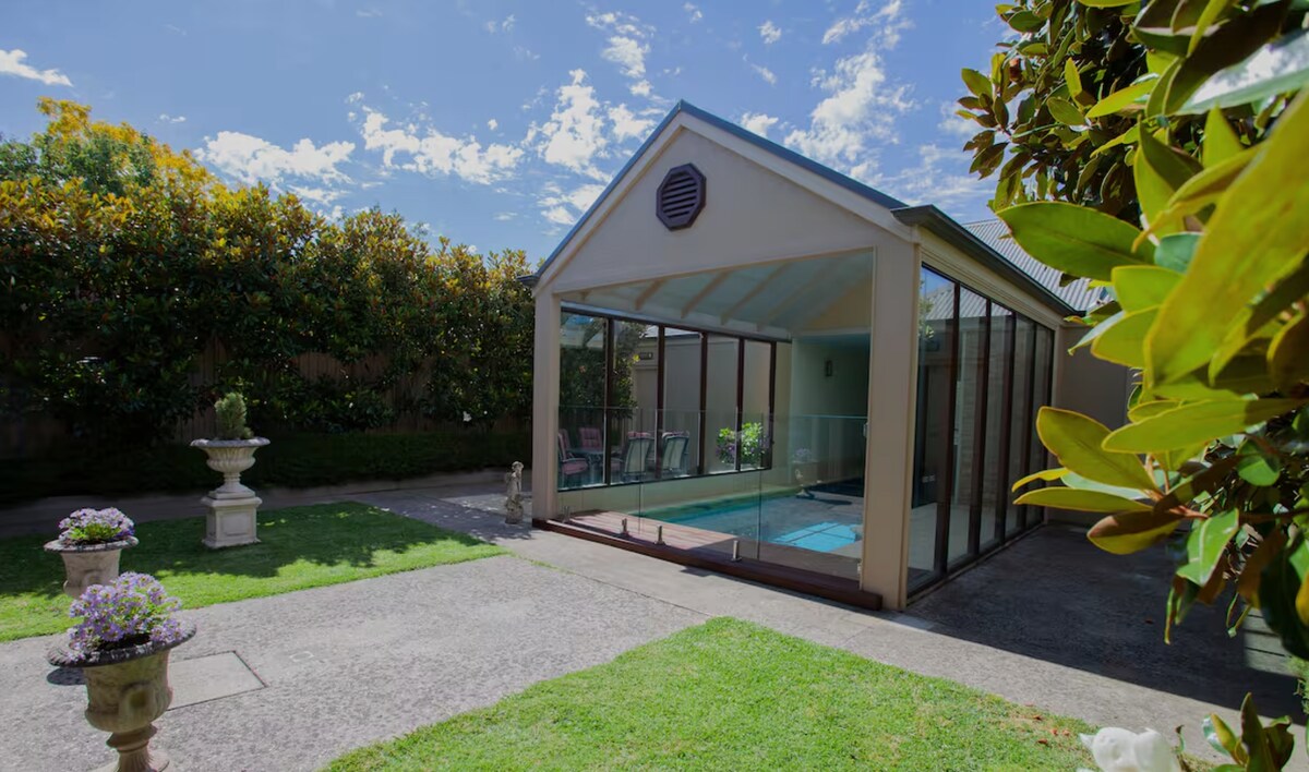 A large, sunlit enclosure surrounds a swimming pool, featuring glass walls and a roof. Lush greenery is seen beyond the enclosure, with well-maintained garden areas and ornamental planters positioned around the edges. The space offers a peaceful retreat for relaxation.