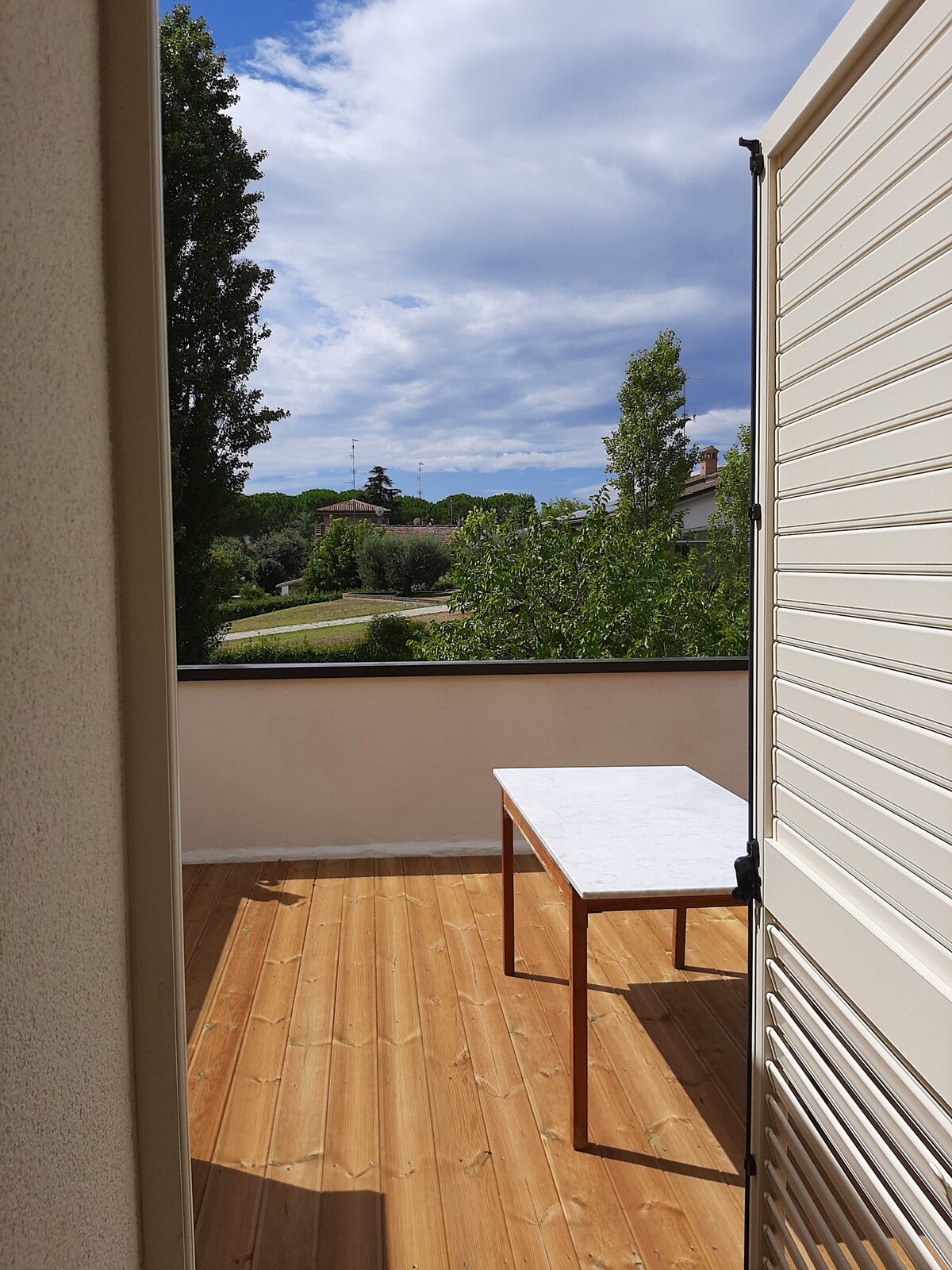 A terrace is featured with a wooden floor and a marble-top table. The entrance is framed by partially open shutters, and a view of greenery and distant hills is visible beyond the railing, enhancing the outdoor space's appeal.