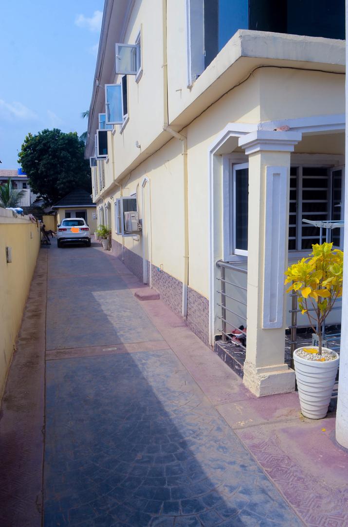 A clean and well-maintained pathway runs alongside the building, bordered by walls and featuring a small potted plant. A parked car is visible at the rear of the home, with nearby windows allowing natural light to enter the space.