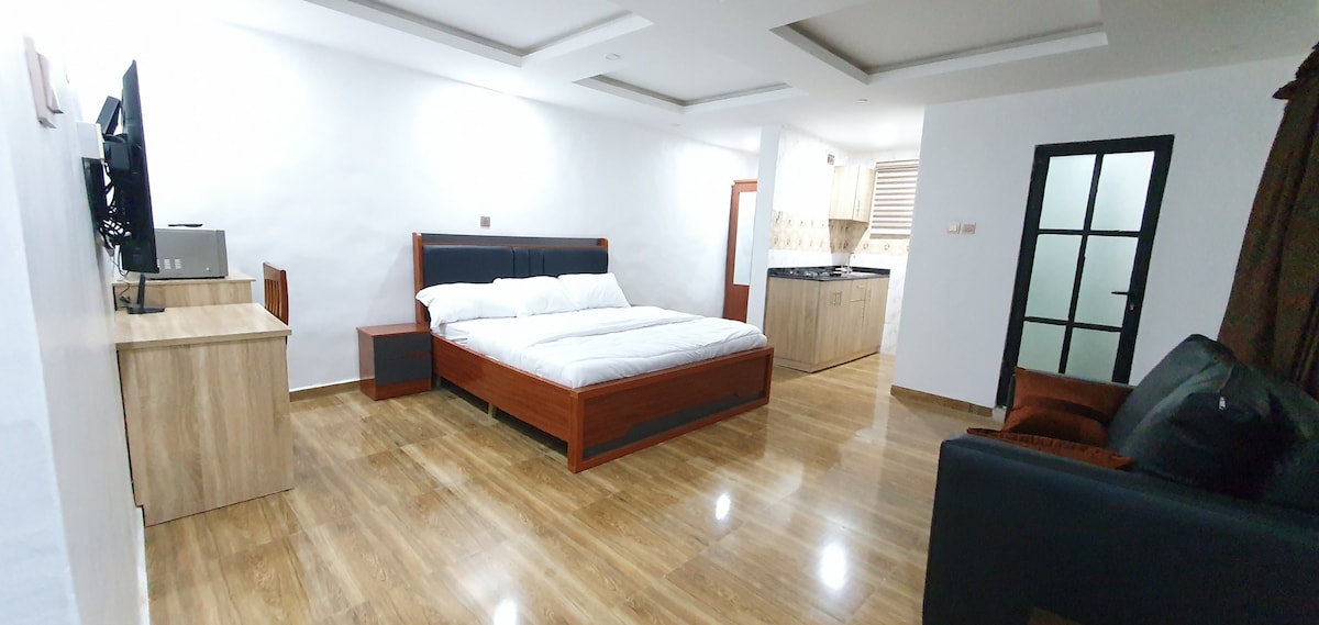 A spacious room features a modern wooden bed with crisp white linens. A comfortable seating area includes a dark couch and a chair. Natural light fills the space, highlighting a flat-screen TV and a wooden dresser. The kitchen area is visible in the background.