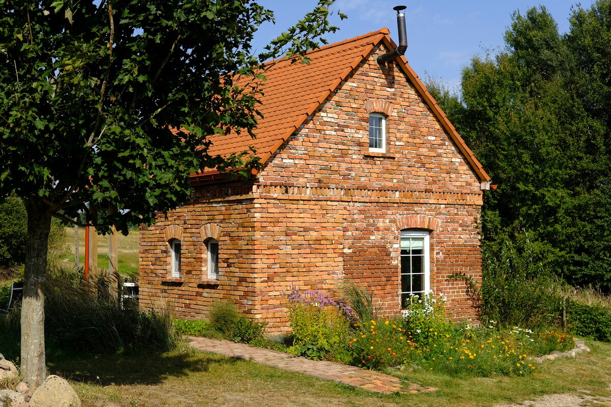 A charming brick cottage with a sloped roof is surrounded by a vibrant garden filled with wildflowers. The structure features several large windows, allowing natural light to enter, and is set against a backdrop of greenery and trees.