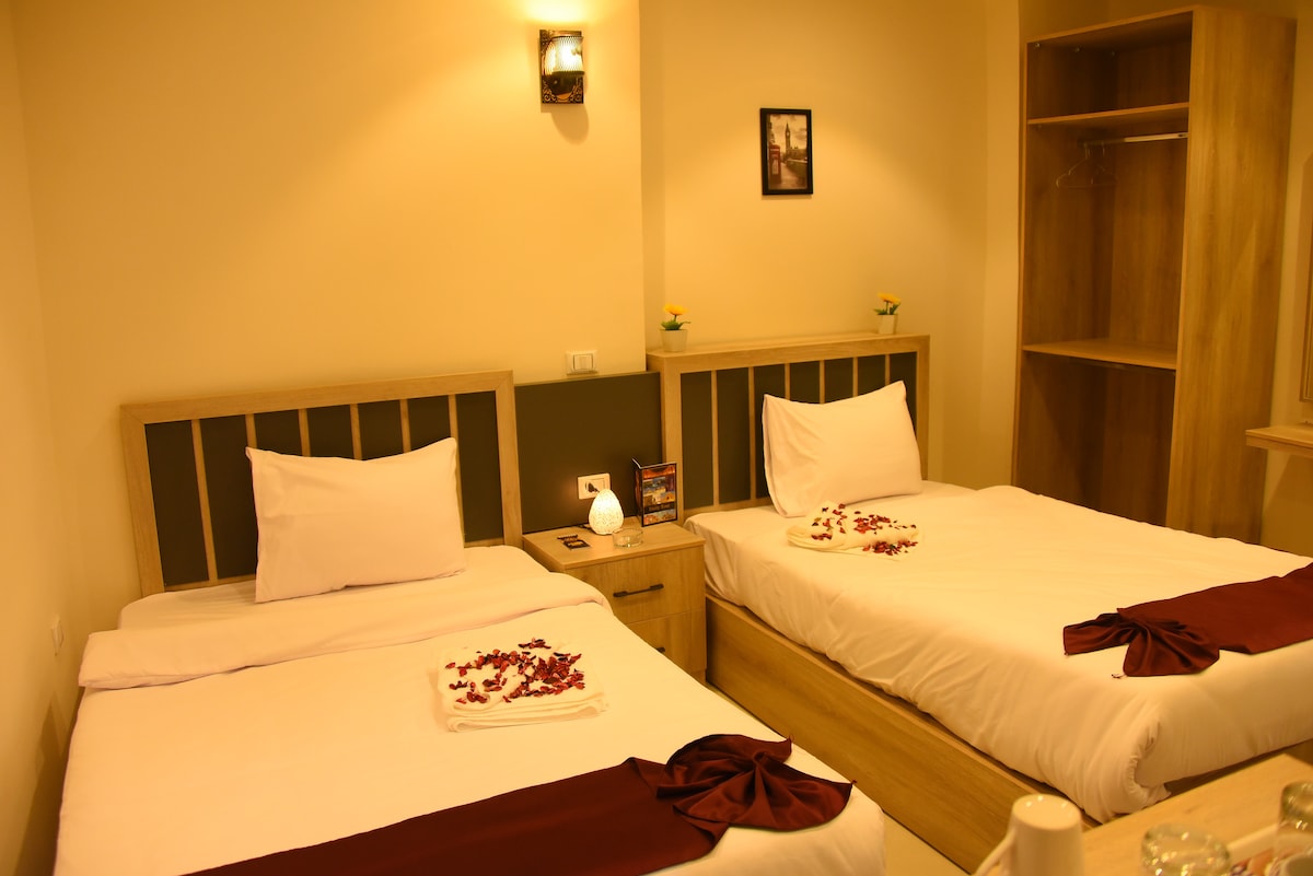 A spacious room featuring two beds adorned with decorative towels and scattered rose petals. Natural light fills the area, highlighting the minimalist wooden furnishings, including a bedside table and a built-in wardrobe. A subtle light fixture adds a warm glow to the atmosphere.