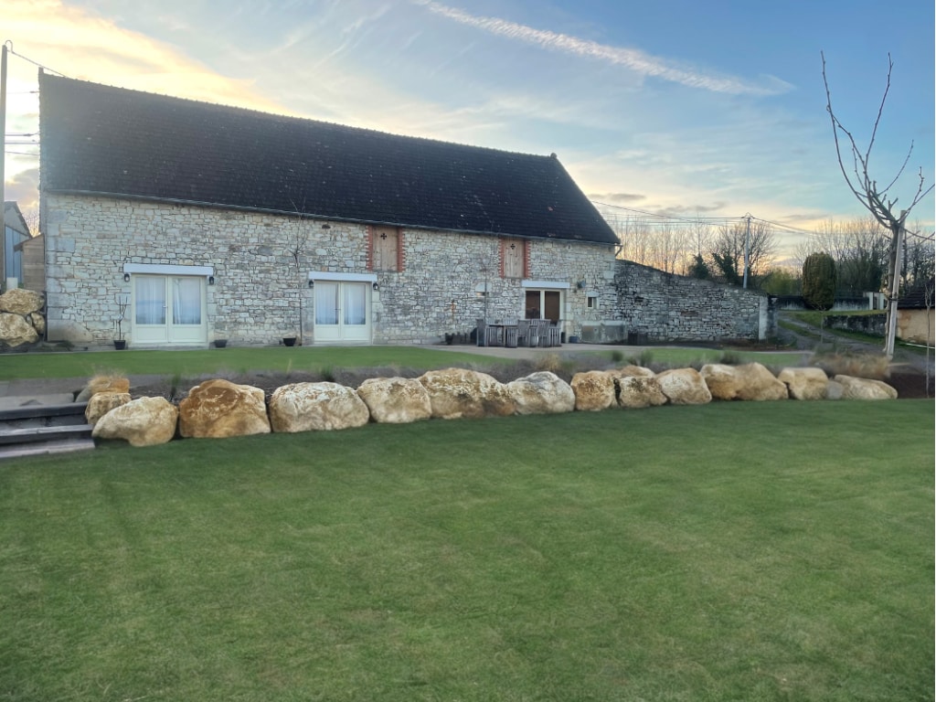 A stone barn with a rustic charm is set against a soft, grassy landscape. Large boulders line the ground in front, complementing the natural surroundings. Light clouds drift across the sky, hinting at a peaceful atmosphere.