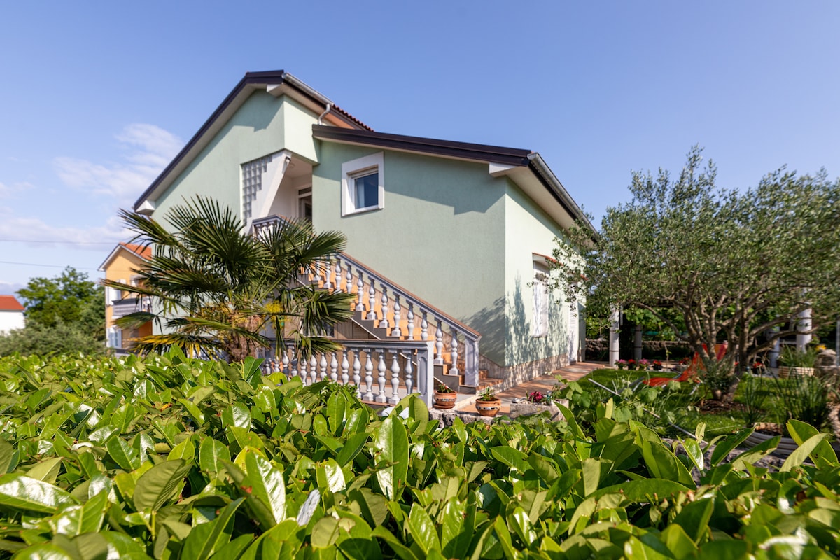 The exterior of a two-story house is presented, featuring a light green facade and a staircase leading to the entrance. Lush greenery surrounds the property, with tropical plants visible at the base, creating a natural and inviting setting.