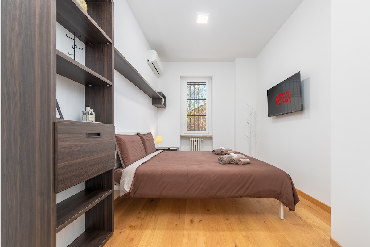 A spacious bedroom features a king-size bed dressed in neutral-toned linens. A Smart TV is mounted on the wall opposite the bed. Natural light enters through a window, and a wooden shelf offers decorative accents. The warm wooden floor enhances the overall ambiance.