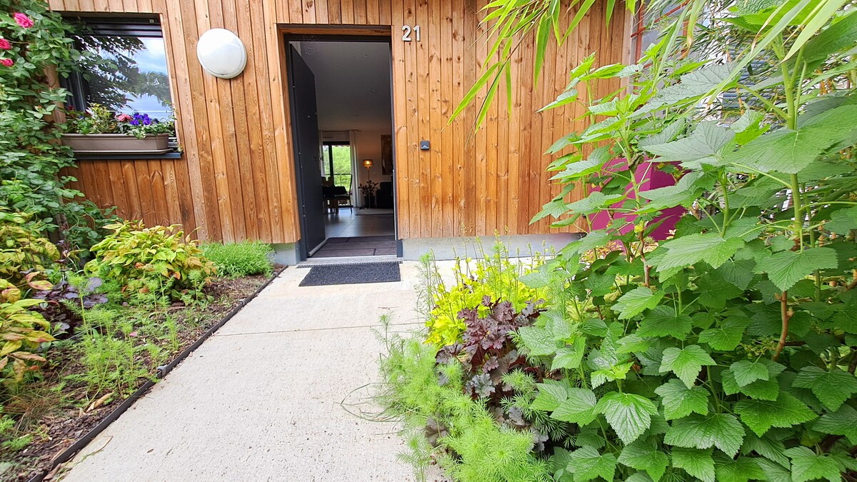 The entrance to the house is framed by vibrant green foliage and colorful flowers, leading to a welcoming open door. A concrete path extends to the entrance, with a black doormat positioned at the threshold.
