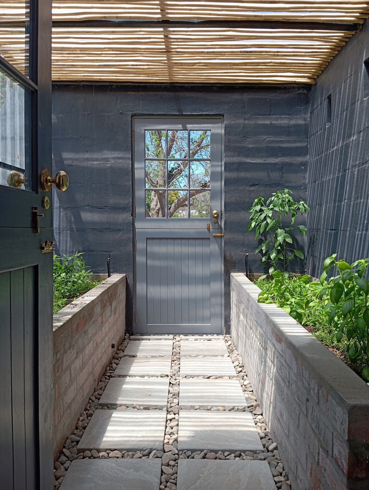 A serene entryway is framed by a gray door and light streaming through a wooden slatted roof. A pathway of stone tiles leads towards the door, bordered by lush greenery in brick planters, creating a tranquil transition between indoor and outdoor spaces.