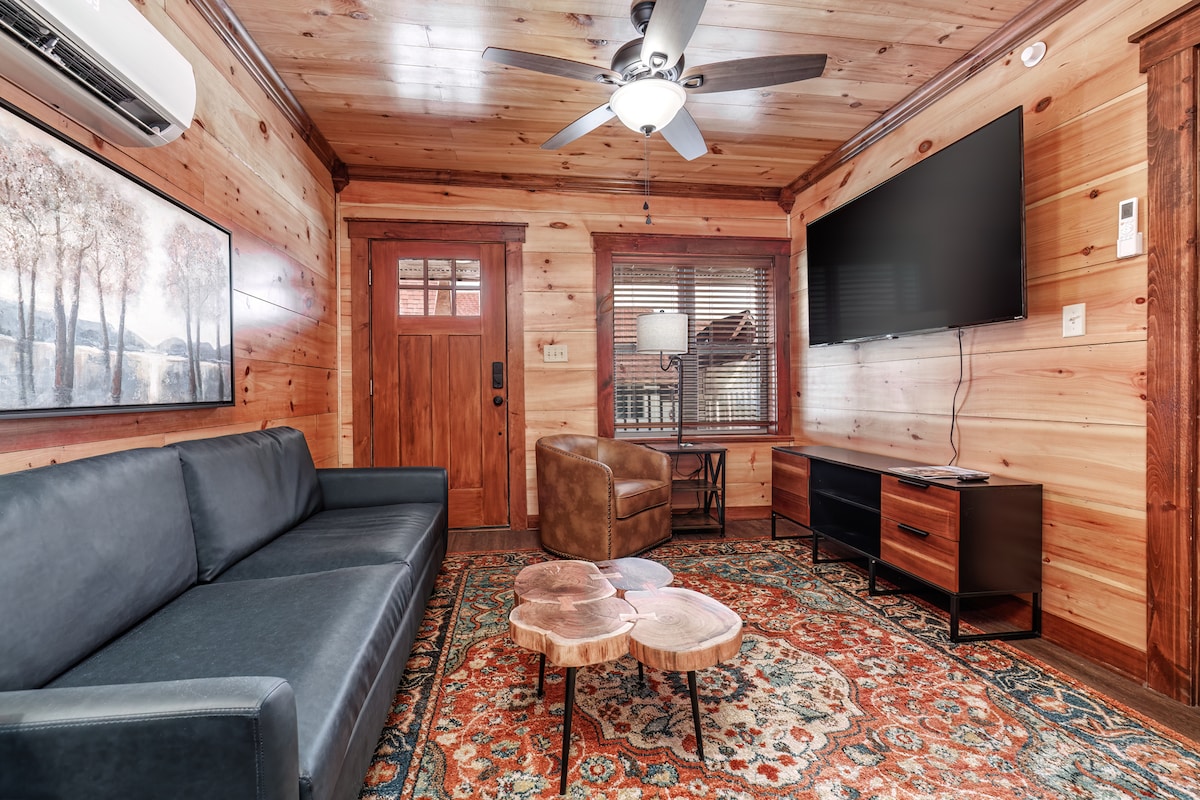 A living area is presented with warm wood paneling, featuring a dark leather sofa and a comfortable armchair. A round wooden coffee table sits center stage, complemented by an area rug with intricate patterns. A flat-screen TV is mounted on the wall, and natural light filters through fitted blinds.