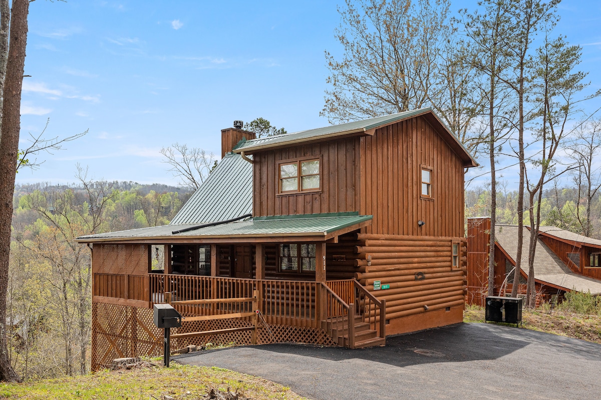 The cabin's exterior showcases a charming wooden structure with a sloped green roof, nestled among the trees. A spacious porch is present, featuring latticework and a barbecue grill, with a paved driveway providing ample parking.
