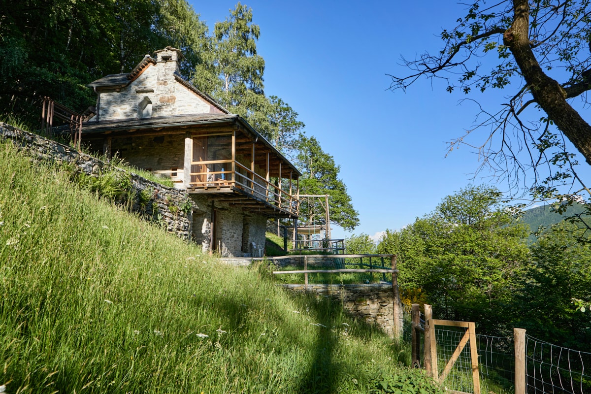 The stone house is set against a backdrop of lush greenery, featuring a spacious balcony that wraps around the south-facing side. A terraced garden area is visible below, bordered by a fence, with trees and foliage enhancing the natural surroundings.