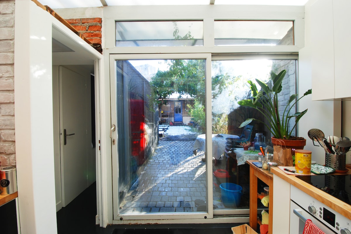 A light-filled kitchen features large glass doors that provide a view of the outdoor patio and garden area. Potted plants are visible on the countertop, while a diverse assortment of dishes and kitchenware is organized nearby.