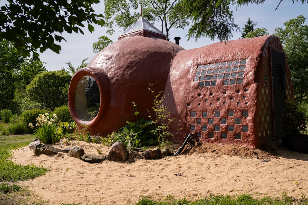 A uniquely shaped dome structure is highlighted in earthy tones, featuring a large round window and decorative glass panes. Surrounded by a landscaped garden, the building blends with nature, showcasing a sandy area and lush greenery that enhances its rustic charm.