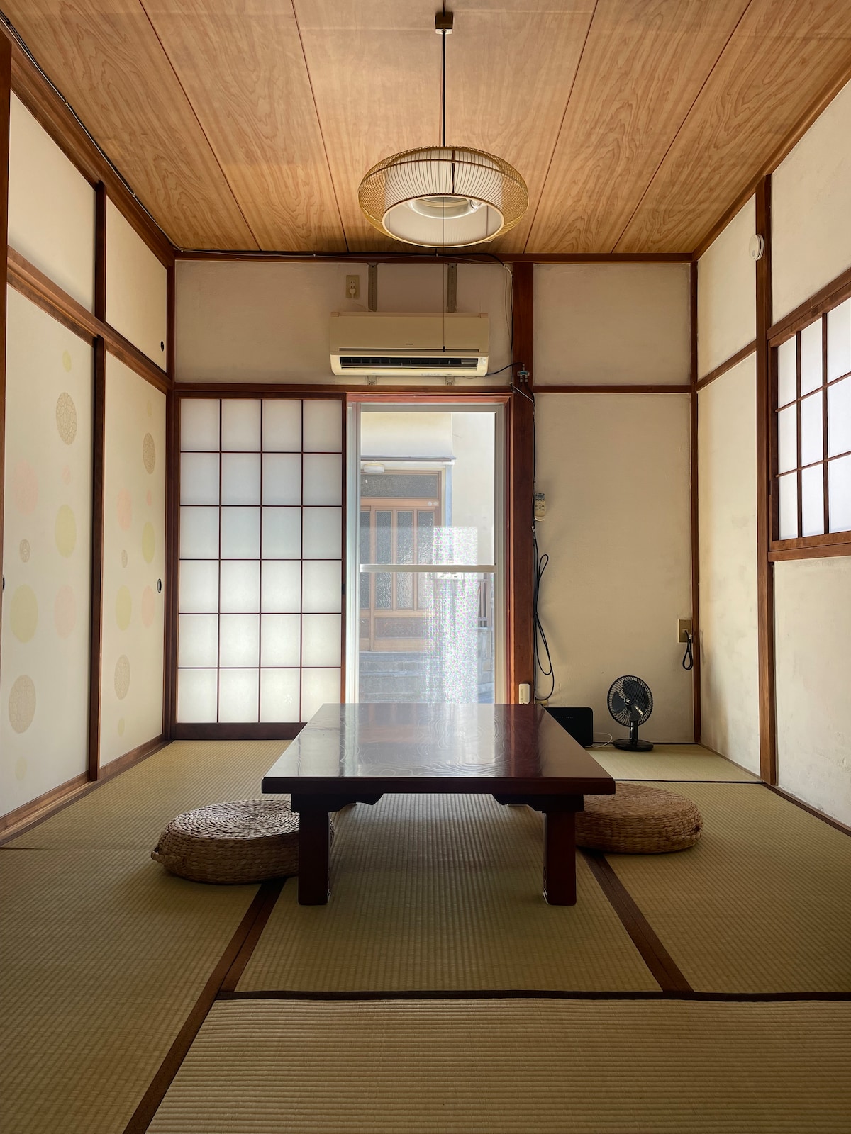 A tatami-walled room features a low table at the center, surrounded by two floor cushions. Light filters through the wooden sliding doors, creating a serene ambiance. An air conditioning unit is mounted on the wall, and a small fan is located on the floor.