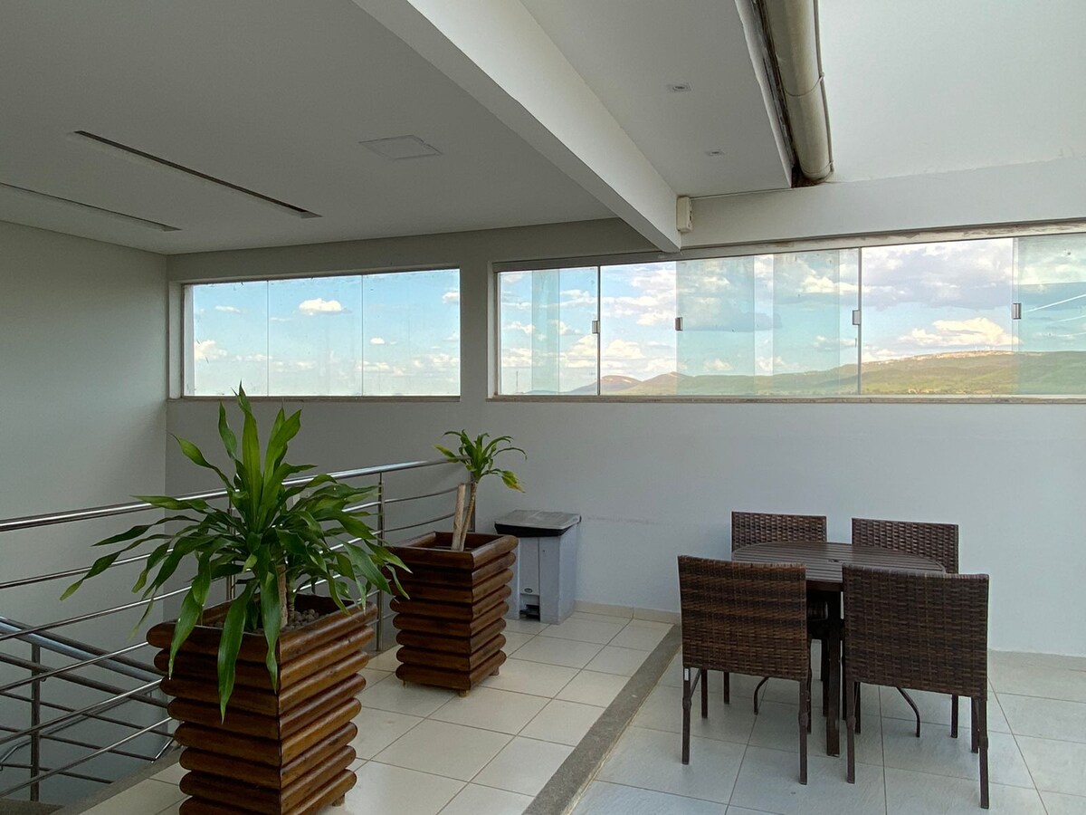 An open space is showcased with large windows allowing natural light to flood in. Two potted plants are positioned prominently, and a small dining table surrounded by four wicker chairs is visible, providing a comfortable area for gatherings. Scenic views can be seen beyond the glass.