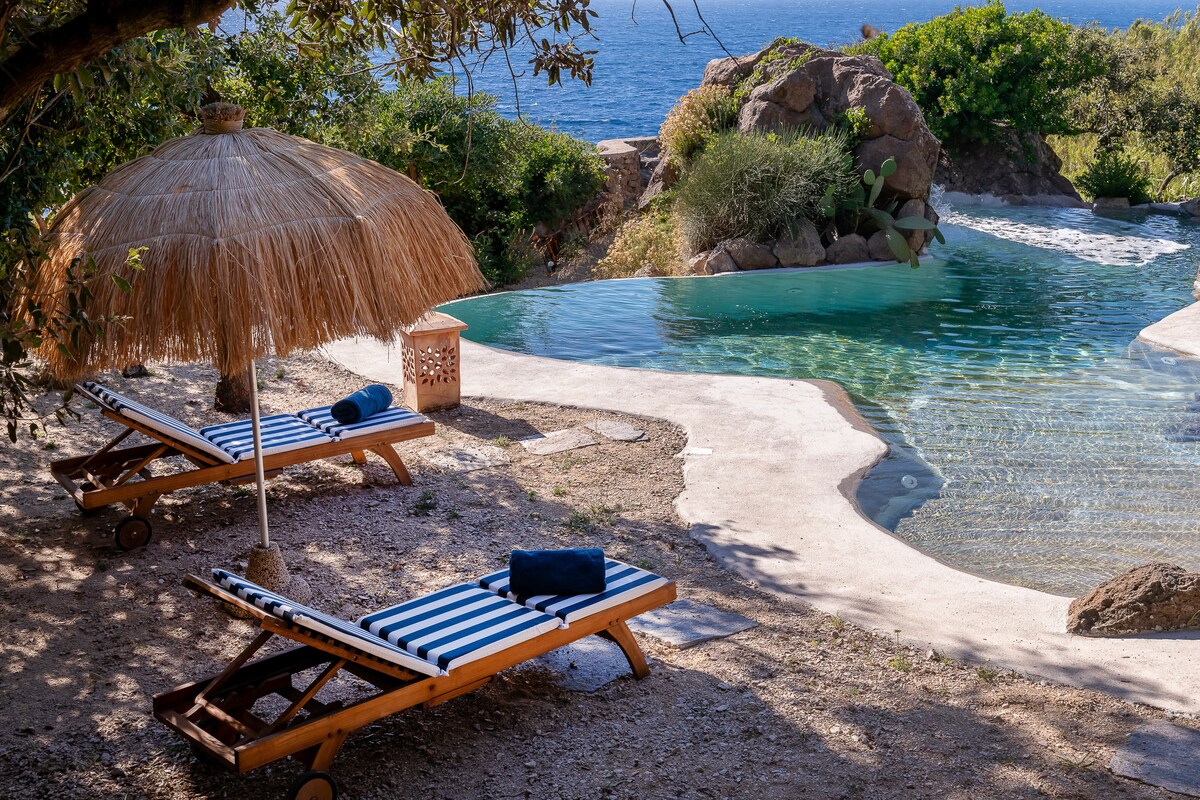 Two sun loungers with striped fabric are positioned under a thatched umbrella by the natural pool. Lush greenery surrounds the area, with smooth rocks forming the pool's edge. The tranquil water reflects the blue of the sea in the background.