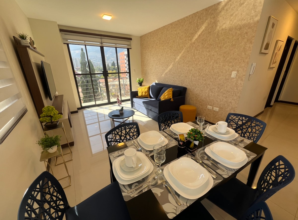 A dining table is set for six, featuring white plates and glasses. A sofa with yellow cushions is positioned next to large windows, offering views of the city. Natural light fills the open living area, enhancing the modern decor.