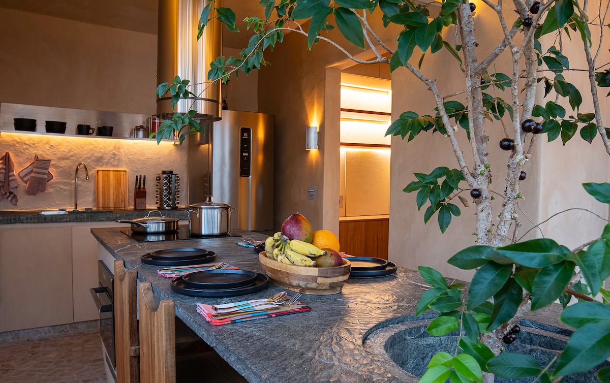 An inviting kitchen features a stone countertop with prepared plates and a bowl of fruit, including bananas and apples. Shelves hold various kitchen utensils. A plant adds a touch of greenery, while modern appliances are visible in a contemporary design.