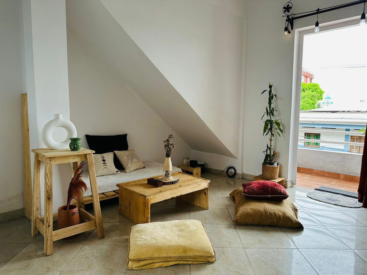 A living area features a minimalist wooden coffee table accompanied by cushions of varying colors on the floor. A small plant sits in the corner, while light enters from a large open door leading to a small balcony.