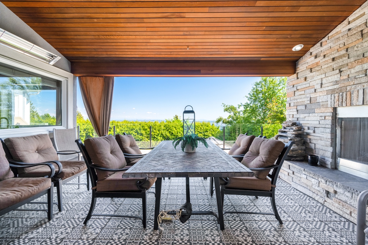An outdoor seating area features a large rectangular table surrounded by several cushioned chairs. Natural stone walls and wooden ceilings complement the space, which is enhanced by scenic views of greenery and distant mountains through large windows.