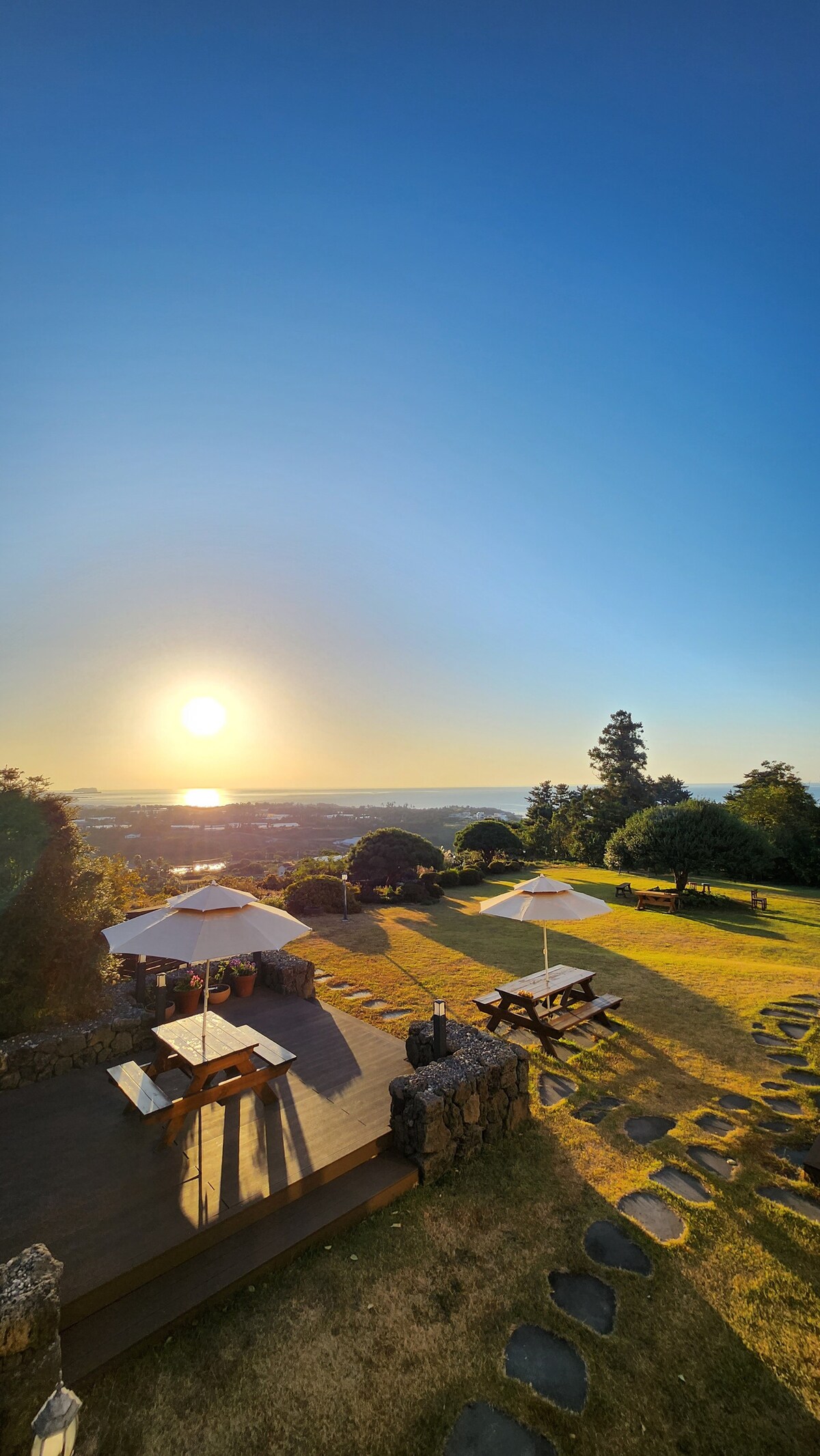 An outdoor seating area with wooden picnic tables is set under umbrellas, positioned on a grassy lawn. The sun is rising over the distant landscape, casting warm light across the scene. Stone pathways wind through the garden, surrounded by lush greenery.