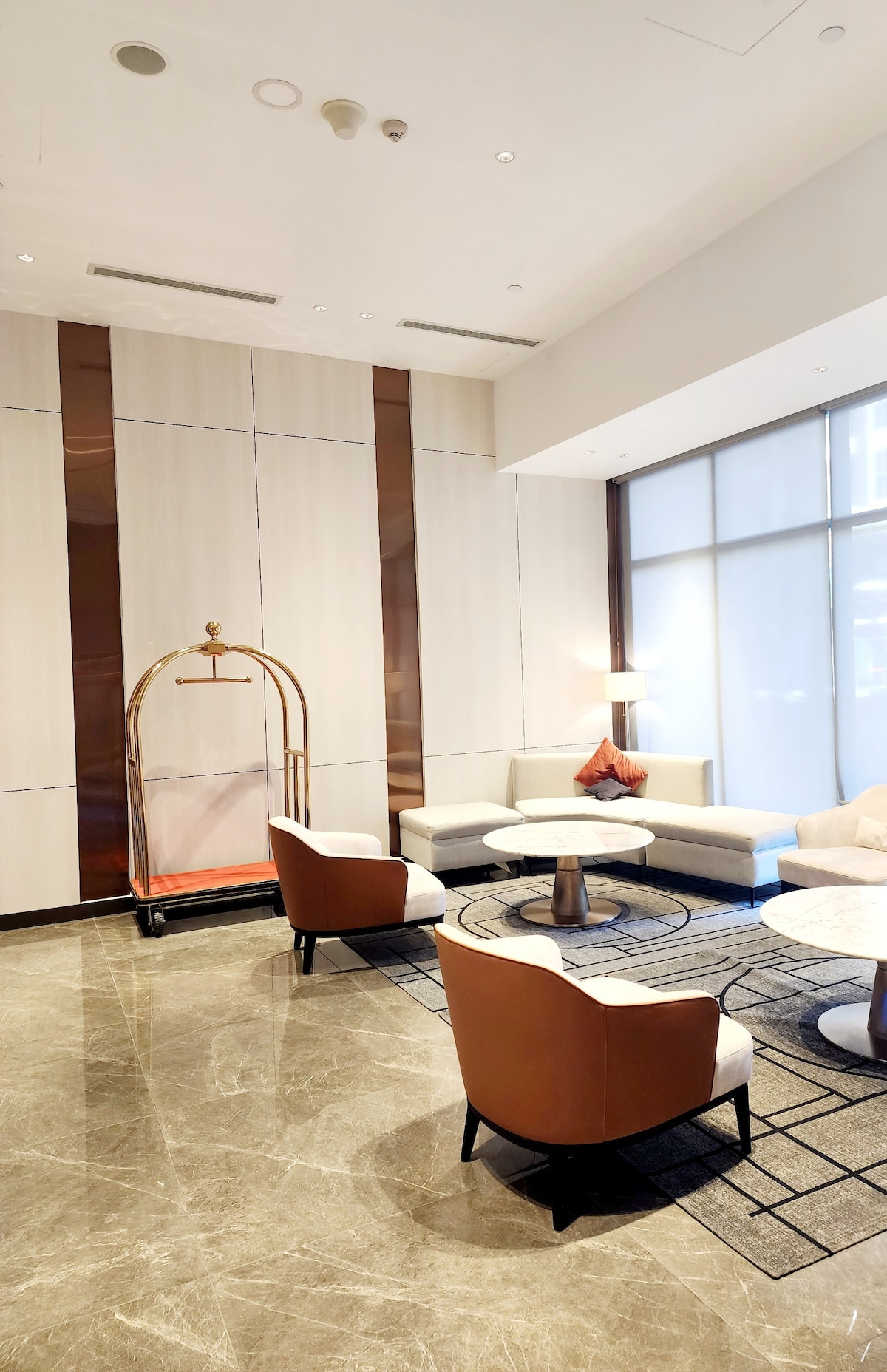 A modern lobby area is highlighted by a light color scheme and sleek design. A curved sectional sofa is positioned around a circular coffee table, complemented by two armchairs. Natural light filters through large windows, enhancing the open atmosphere.