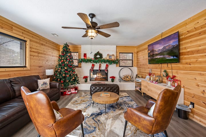 Festive and cozy living room during the holidays with warm wood walls, leather seating (queen sofa bed), electric fireplace, plenty of seating, and a large TV. Natural light and a ceiling fan make it perfect for relaxing or gathering.