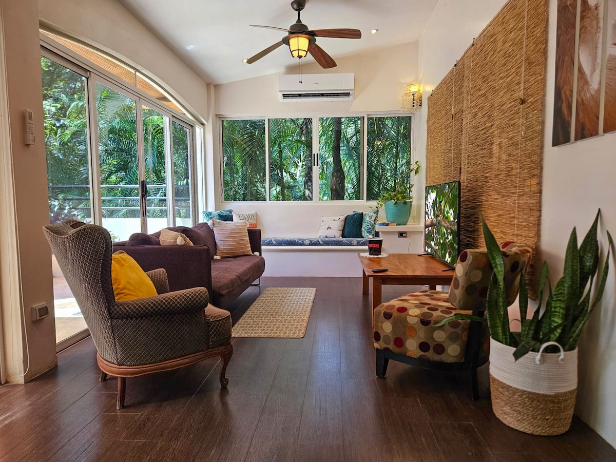 A cozy living area is depicted, featuring a brown sofa and a patterned chair. Natural light floods through large windows overlooking lush greenery. A flat-screen TV is positioned on a wooden table, and a comfortable window seat offers additional lounging space.
