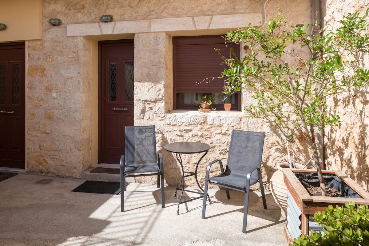 A communal courtyard is depicted, featuring two metal chairs positioned around a small round table. Natural stone walls create a rustic backdrop, with a decorative plant in a planter nearby. Entryways with wooden doors provide access to the private studio.