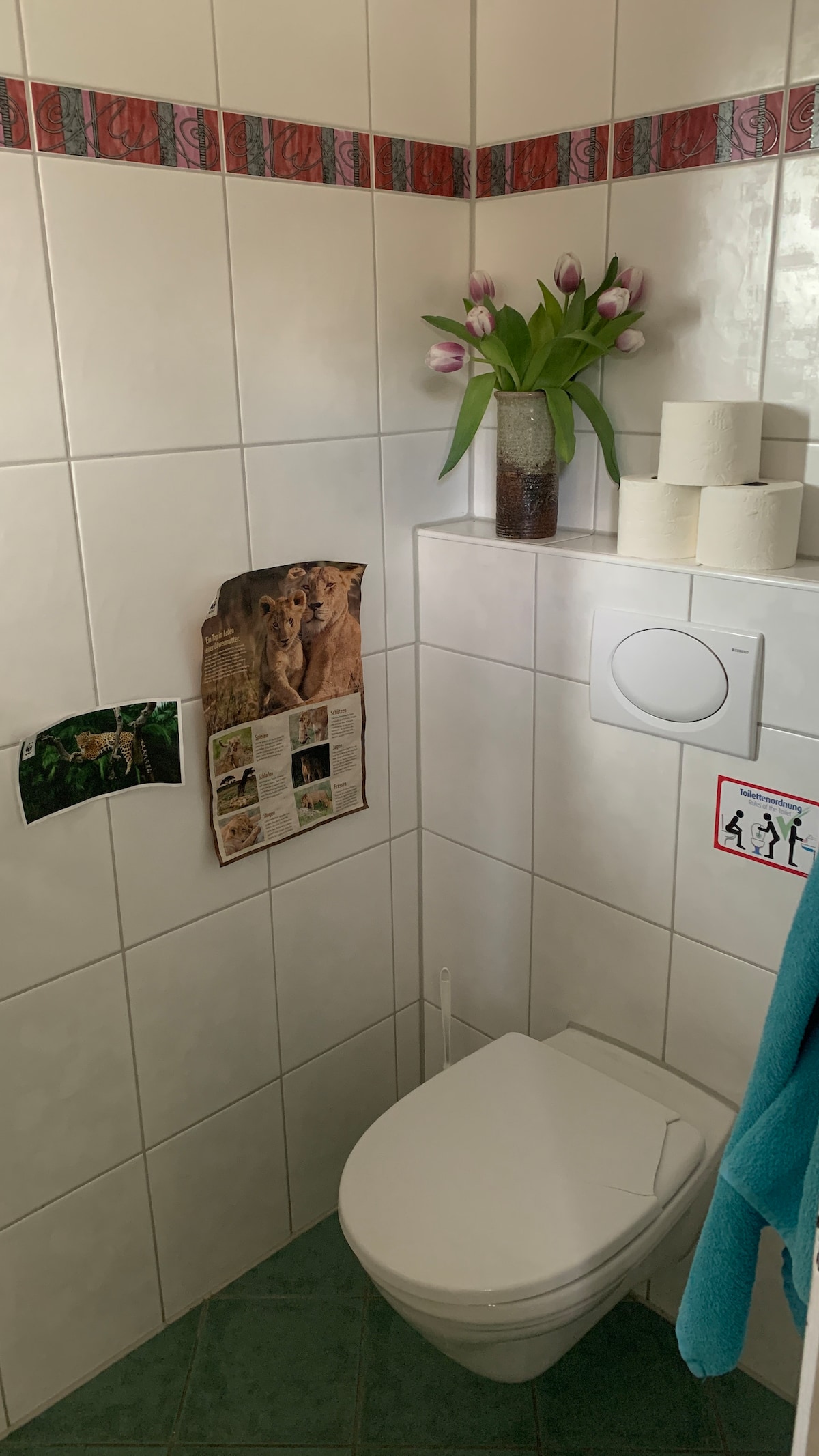 The bathroom features white tiled walls with decorative trim. A modern toilet is positioned beside a potted arrangement of tulips. Three rolls of toilet paper are neatly arranged on a shelf, and a small, colorful towel hangs nearby. Artwork is displayed on the wall.
