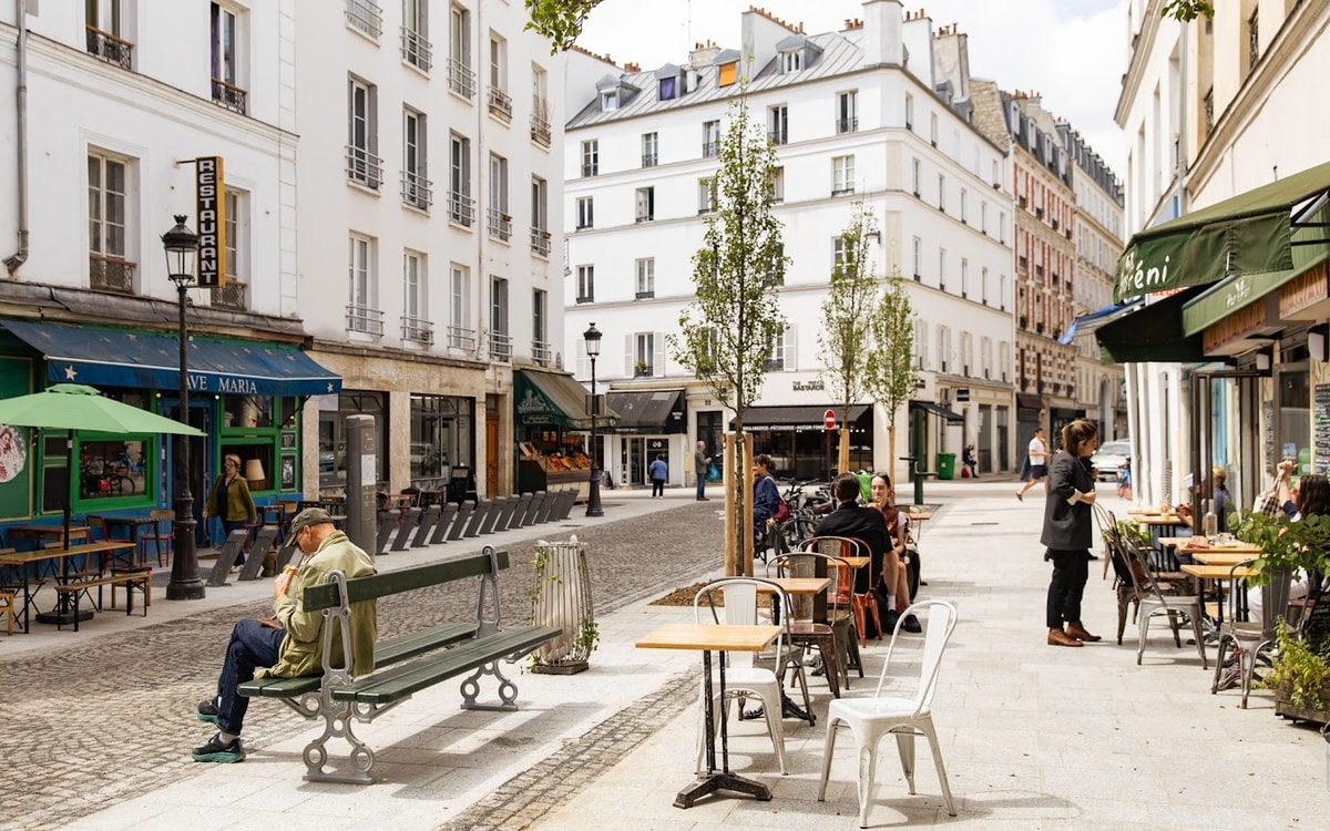 A lively street scene depicts a charming area with outdoor seating and people enjoying the ambiance. Trees provide greenery along the pedestrian walkway, while café tables invite guests to relax. The architectural details of the surrounding buildings showcase traditional Parisian design elements.