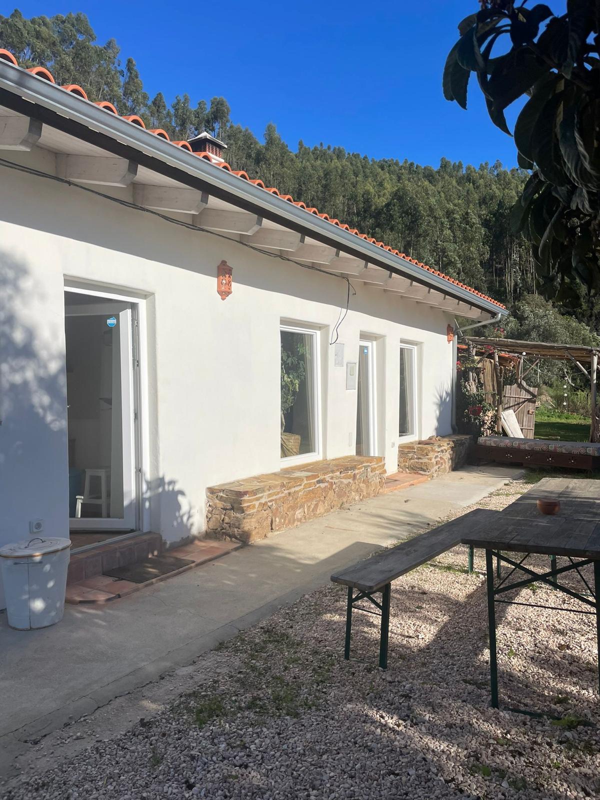A single-story building features a stone base and large windows that invite natural light. The rustic outdoor area is complemented by a long, simple table surrounded by gravel. Lush green trees serve as a backdrop, enhancing the connection to nature.