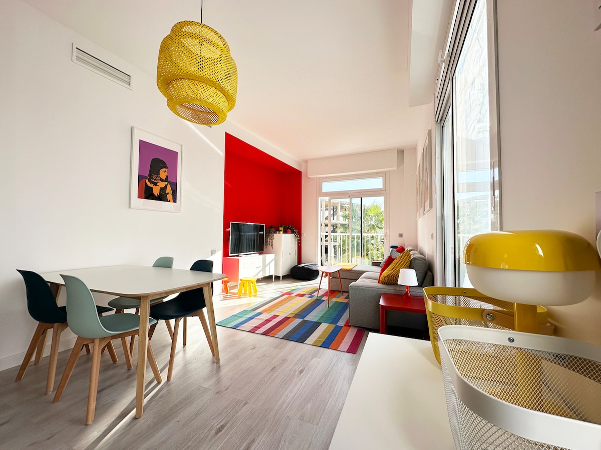 A bright living area features a colorful area rug and a mix of modern furnishings. A spacious dining table with six chairs is complemented by a sofa and wall-mounted TV. Large windows allow natural light to fill the room, creating a cheerful atmosphere.