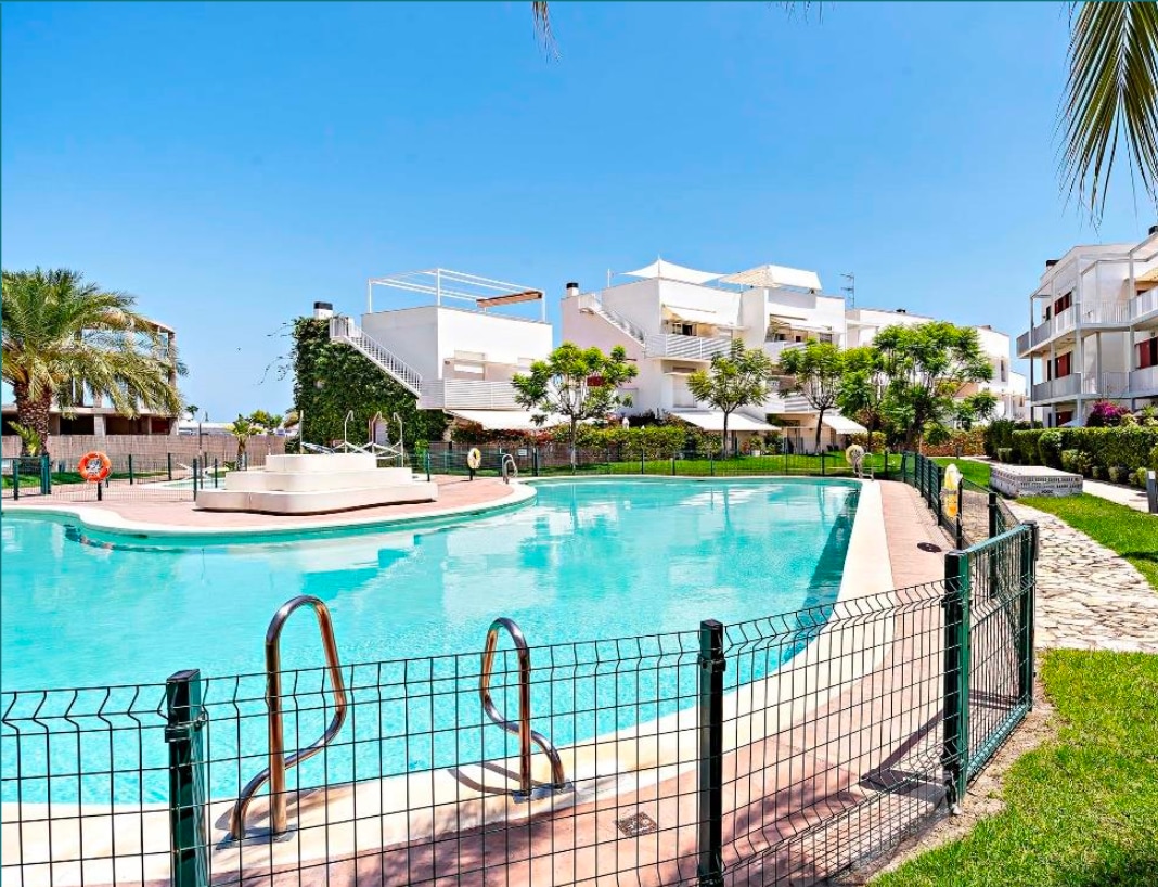 A clear view of the communal swimming pool is presented, surrounded by manicured gardens and lounge areas. The pool features a gentle slope and is bordered by a secure fence. Sun loungers are situated nearby, inviting relaxation under the sunny sky.