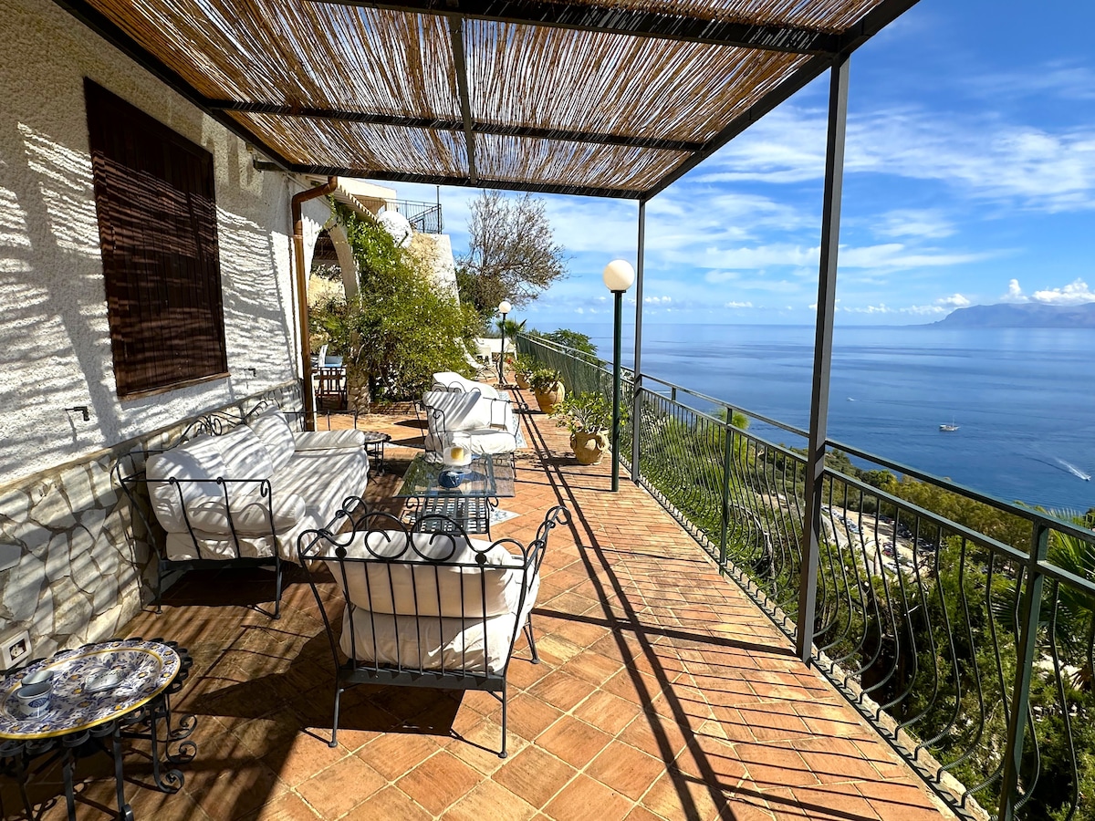 An outdoor terrace is showcased, featuring a shaded seating area with comfortable sofas and a glass table. The expansive view of the sea and coastline is visible in the background, complemented by a clear blue sky with a few scattered clouds.