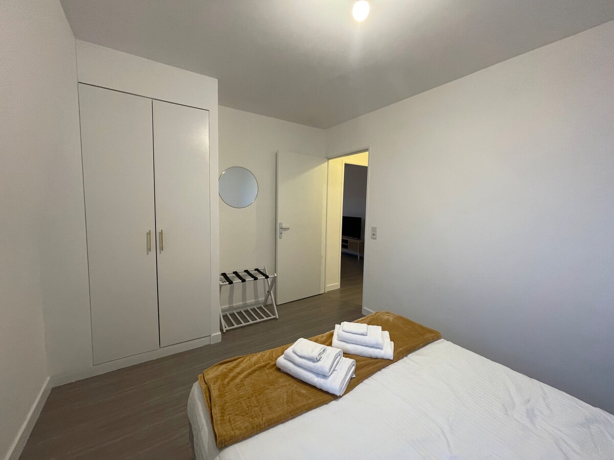 A well-lit bedroom is presented, featuring a spacious bed dressed with warm throw blankets and neatly arranged towels. A round mirror is mounted on the wall, and a closet with sliding doors is visible. A door leads to an adjoining room, enhancing the inviting atmosphere.