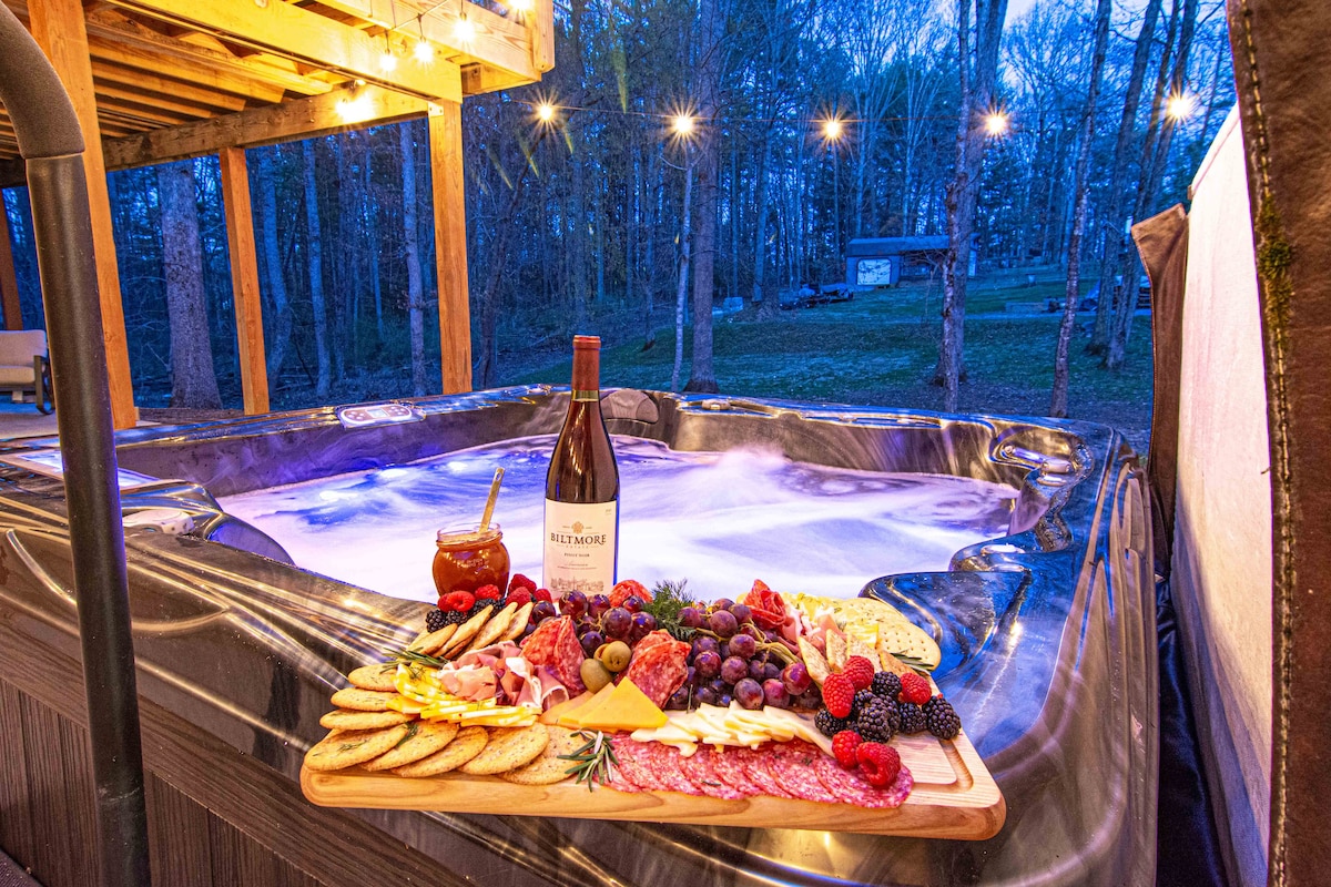 A luxurious hot tub is positioned on a spacious deck, surrounded by trees and illuminated by soft string lights. A wooden platter is filled with an assortment of cheeses, cured meats, crackers, and fresh fruit, complemented by a bottle of wine and a cocktail, enhancing the outdoor experience.