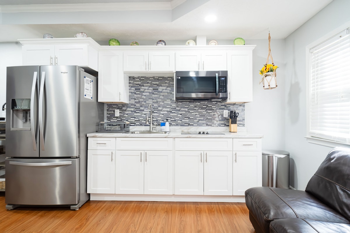 A modern kitchen features stainless steel appliances, including a refrigerator and microwave. Light-colored cabinetry contrasts with a stylish backsplash. A sink is positioned between the appliances, while counter space is available for meal preparation. Natural light streams in from a nearby window.