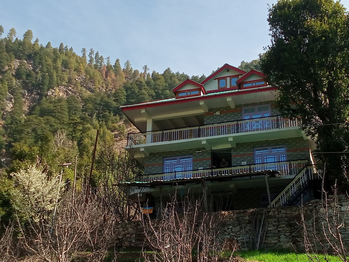 A three-story building with a stone facade is set amidst a backdrop of lush green hills. The structure features multiple balconies with railings, offering a view of the surrounding landscape. The property is bordered by budding apple trees, adding to the natural charm of the location.