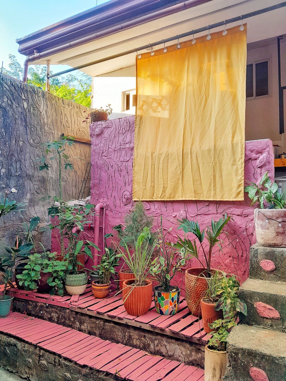A colorful outdoor space features a set of terracotta pots filled with various greenery along a pink-striped walkway. A yellow curtain hangs above a textured pink wall, creating a vibrant backdrop for the plants. Steps lead down, enhancing the cozy ambiance.