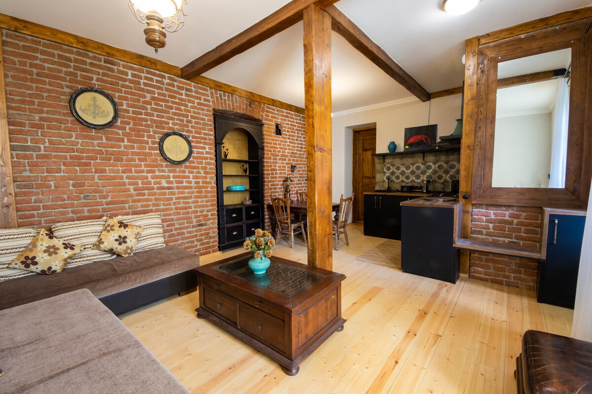 Exposed brick walls define the living area, which includes a cozy seating arrangement with two couches adorned with decorative pillows. A wooden coffee table is centered in the room, complemented by rustic shelves and accent pieces. The kitchen area features dark cabinetry and a stylish backsplash.