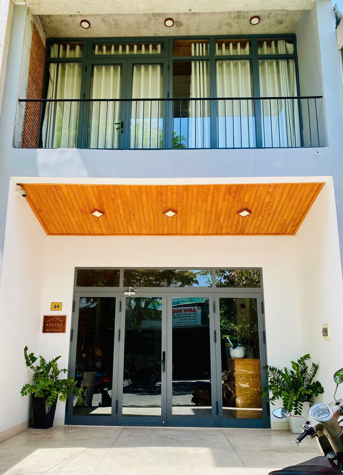 The exterior of the apartment is characterized by a modern facade with large glass doors framed by plants. Above, spacious balconies with light curtains allow natural light to enter, while warm wooden accents on the ceiling add a welcoming touch.
