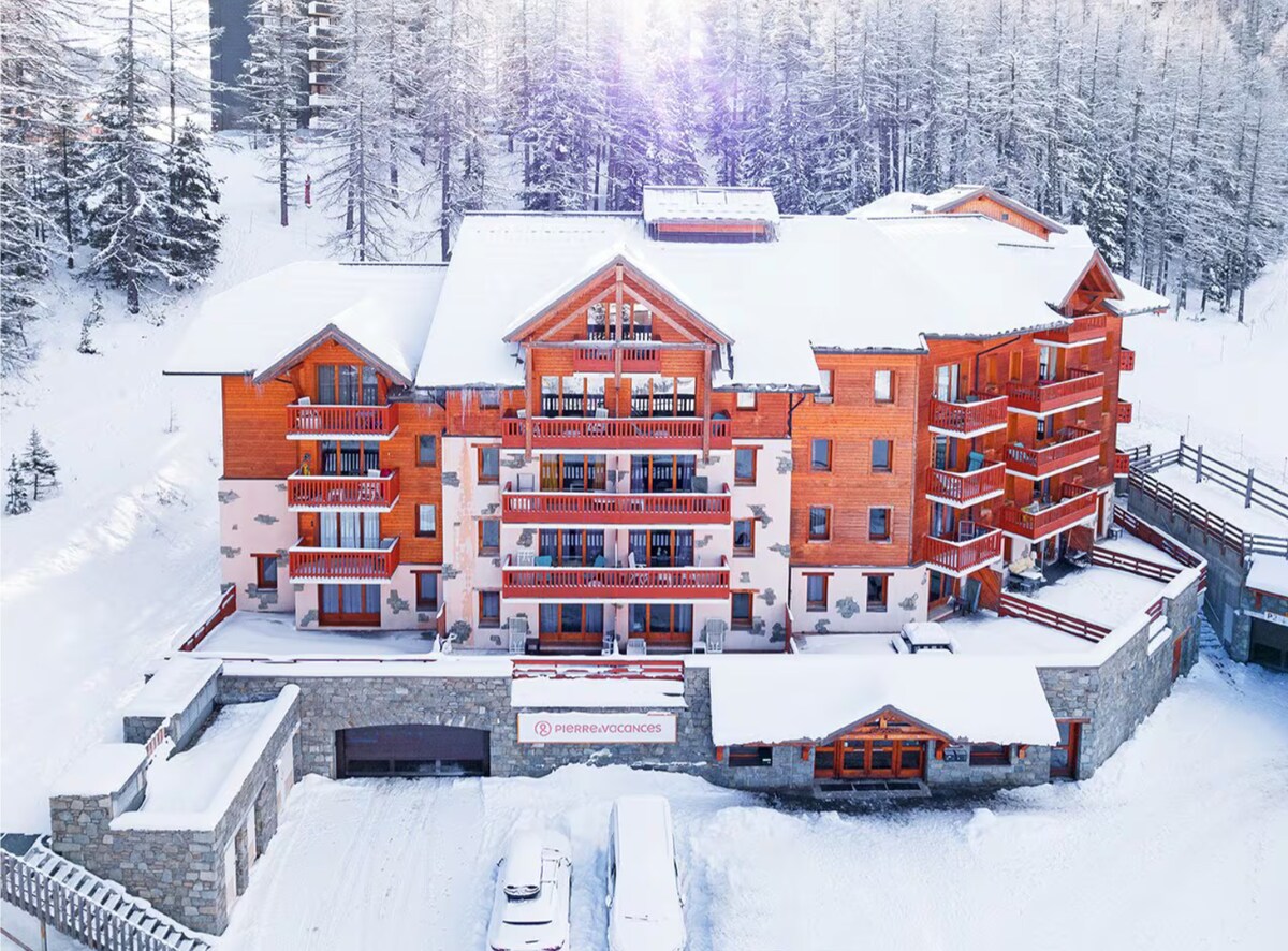 An aerial view of a spacious wooden lodging surrounded by snow-covered trees. The building features multiple balconies, each adorned with wooden railings. A covered parking area is visible at the front, alongside a clear pathway leading towards the entrance.