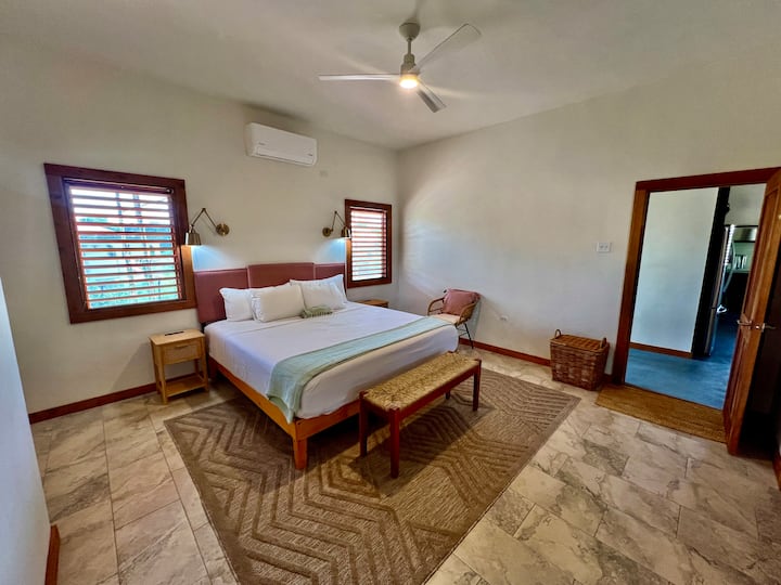 【ベッドルーム１】専用バスルーム付きのメインレベルのキングベッドルーム。 エアコンと天井ファン。 フレンチドアからバックベランダとプールにアクセスできます。 メインキッチンと屋内リビングエリアから離れた場所にあります。 