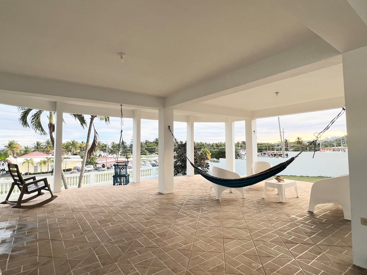 A spacious covered patio is showcased, featuring a hammock and a wooden rocking chair. The floor is tiled in a neutral tone, and large columns support the open structure. A view of the marina is visible in the background, framed by palm trees.