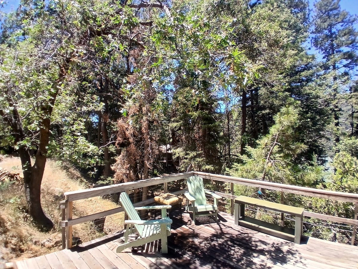 A wooden deck is surrounded by lush trees, providing a natural setting. Two light green Adirondack chairs are positioned for relaxation, with a small table nearby. Sunlight filters through the leaves, creating dappled shadows on the deck surface.