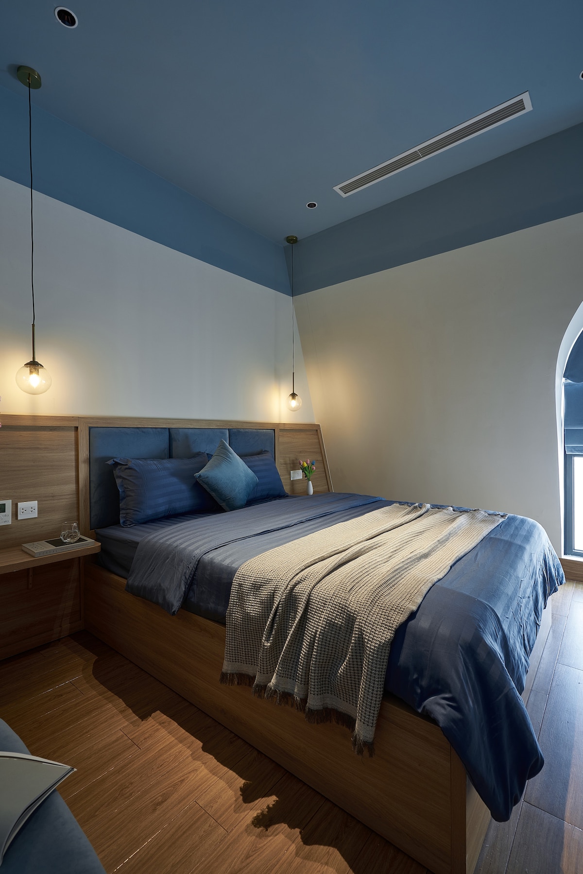 A serene sleeping area features a wooden bed frame dressed in deep blue linens and a soft throw blanket. Ambient lighting is provided by two pendant lights above. A bedside table holds a small plant, complementing the calming color scheme and natural materials.