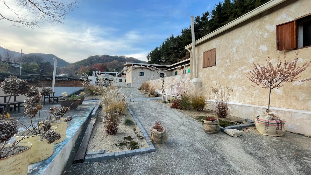 A pathway is bordered by decorative pots and natural plantings, leading to a series of buildings. The scenic backdrop features a hillside covered in trees, under a softly lit sky.
