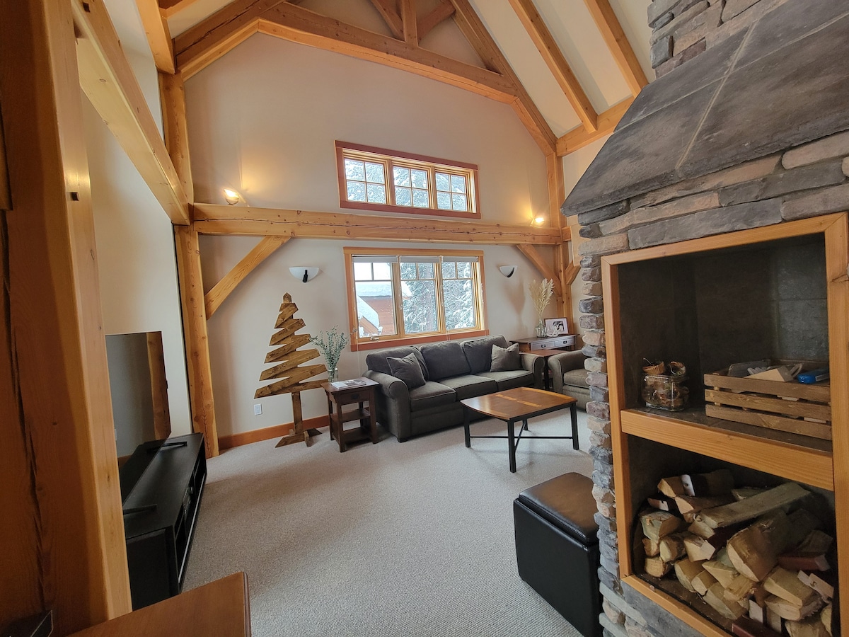 An open and inviting living area features high wooden beams and large windows that allow natural light to fill the space. A cozy seating arrangement faces a double-sided stone fireplace, with a decorative wooden tree positioned in the corner. A stack of firewood is visible beside the fireplace.