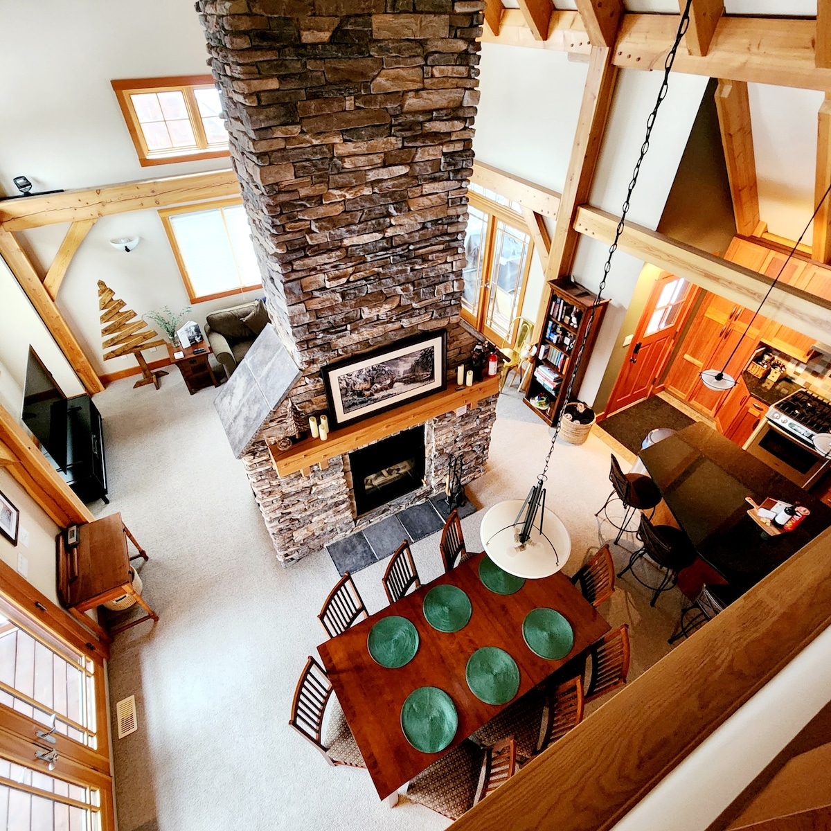 The open-concept main floor is highlighted by a towering double-sided stone fireplace, surrounded by a tasteful dining area with a wooden table set for eight. Light floods in through large windows, illuminating the rustic charm of timber framing and cozy furnishings throughout the space.
