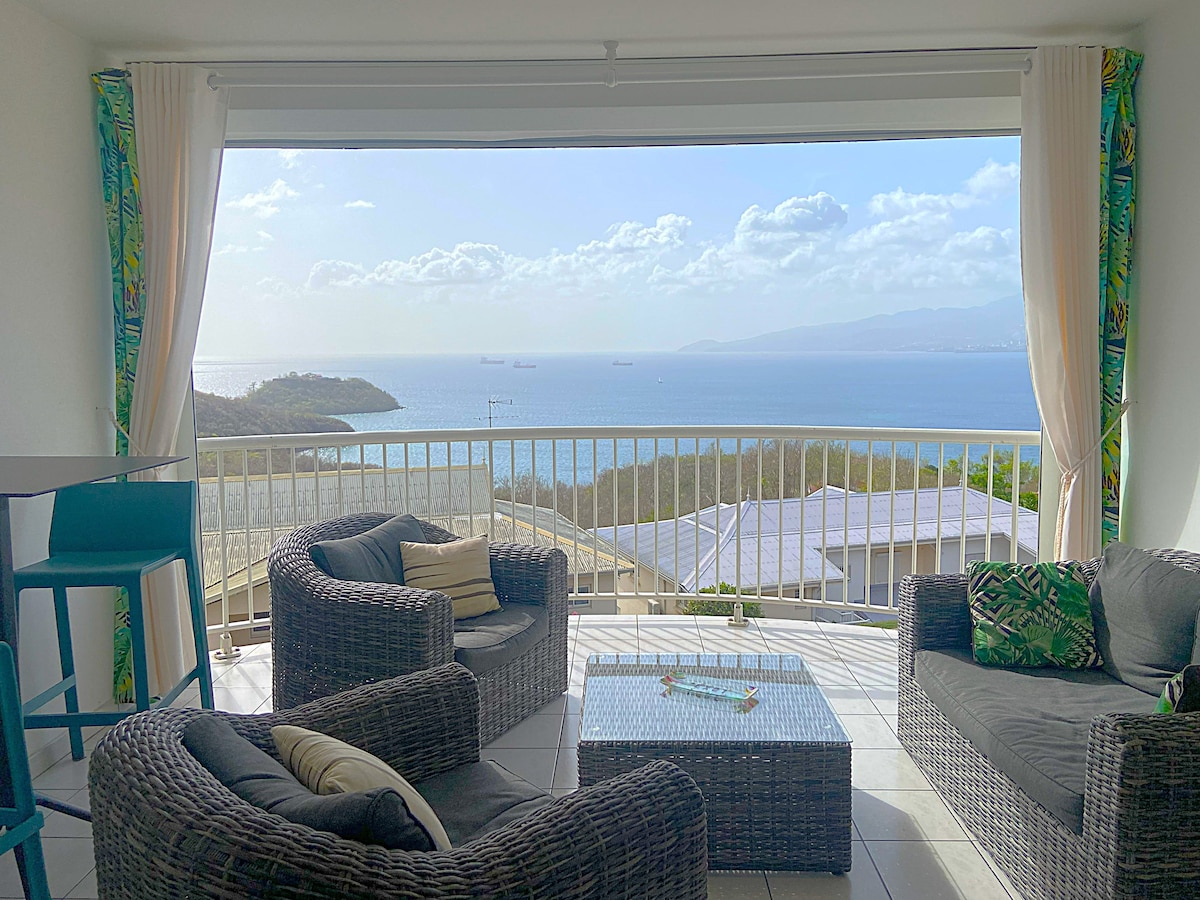 An open terrace presents a panoramic view of the Caribbean Sea, featuring comfortable seating arrangements made of dark wicker. Two chairs and a table are situated under flowing curtains, allowing natural light to illuminate the space while maintaining a serene atmosphere.